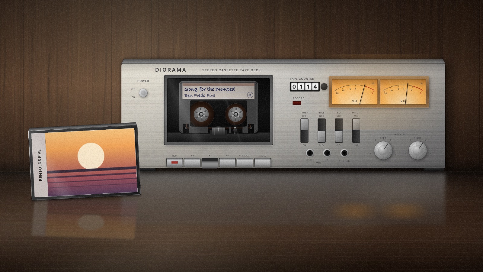 A cassette deck standing on a polished walnut table against a panelled wall, its VU meters lit, the track title hand-written on the tape's label.
